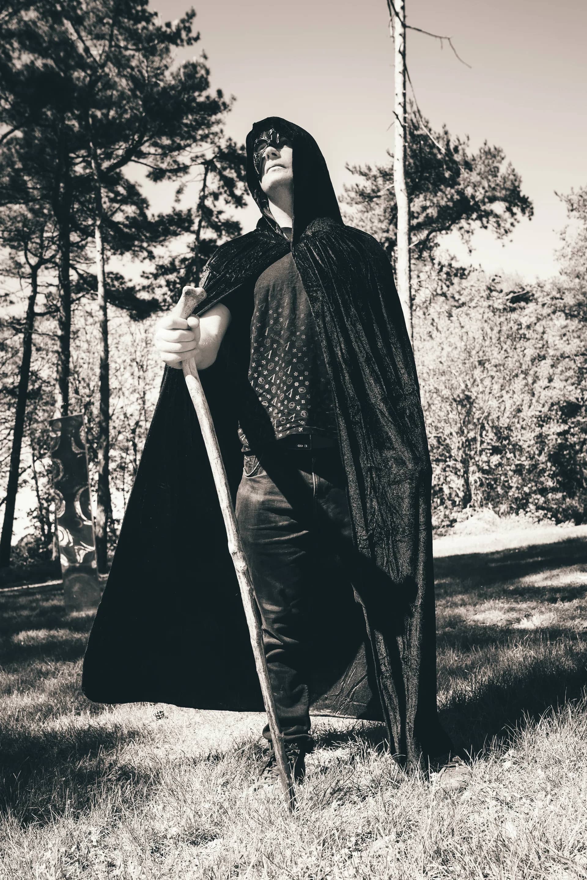 A hooded figure in a long black cloak standing among the pines, one hand on a tall wooden staff, the frame in near monochrome.