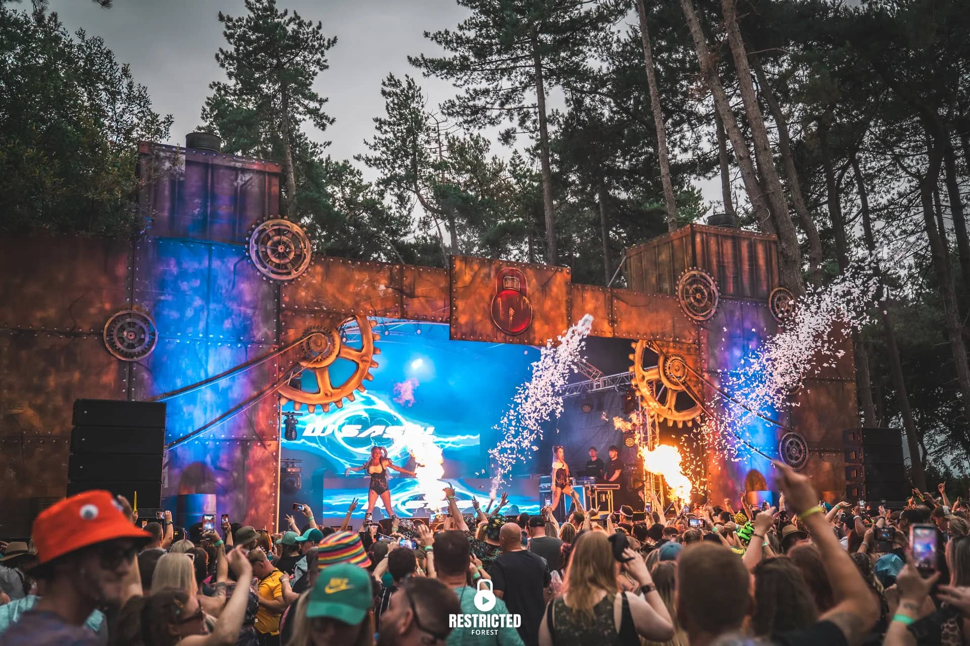 Steampunk cog stage with pyro firing in daylight, crowd filming on phones.