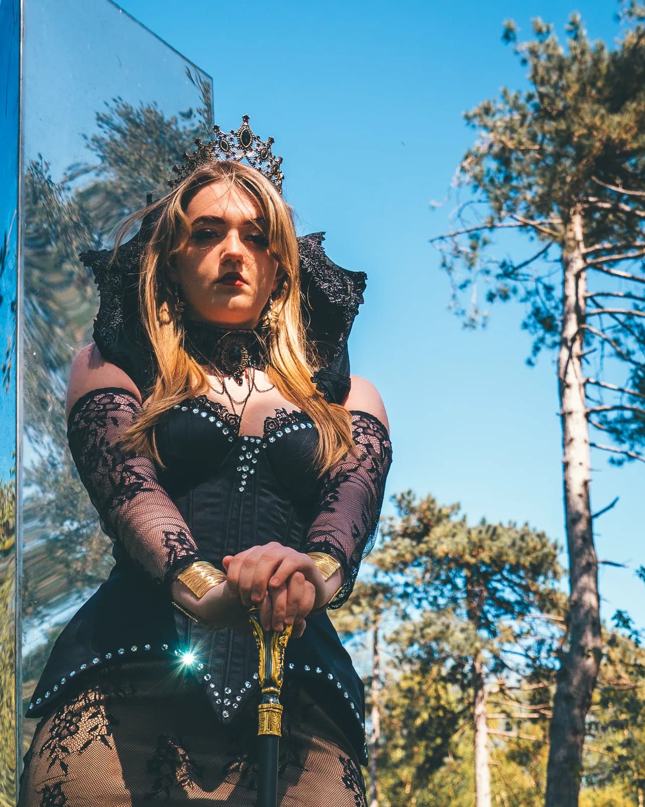 The Empress, guardian of the Castle Realm: a dark crown, a black corseted gown and gold gauntlets on a staff, photographed in the pines.