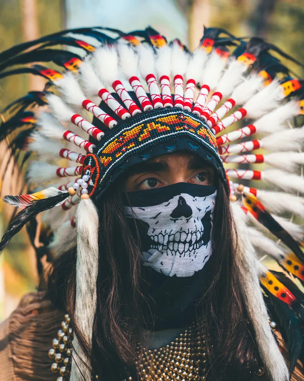 The Oracle, guardian of the Pyramid Realm: a feathered headdress and a skull-painted face covering, photographed in the pines.