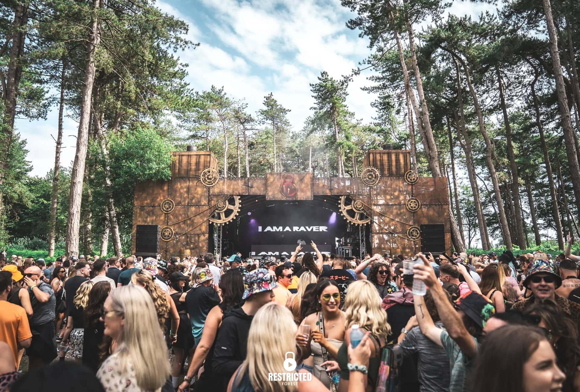 Main stage in daylight with I Am A Raver on the screen, crowd filling the clearing.