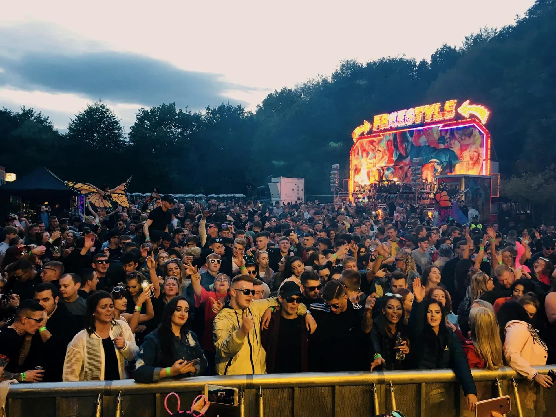 Crowd packed along the front barrier at dusk, a lit fairground ride and the treeline behind them