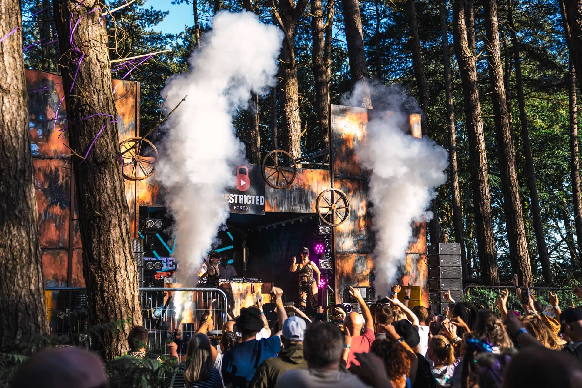 The cog stage with white smoke jets firing past the pines, the crowd with arms raised below.