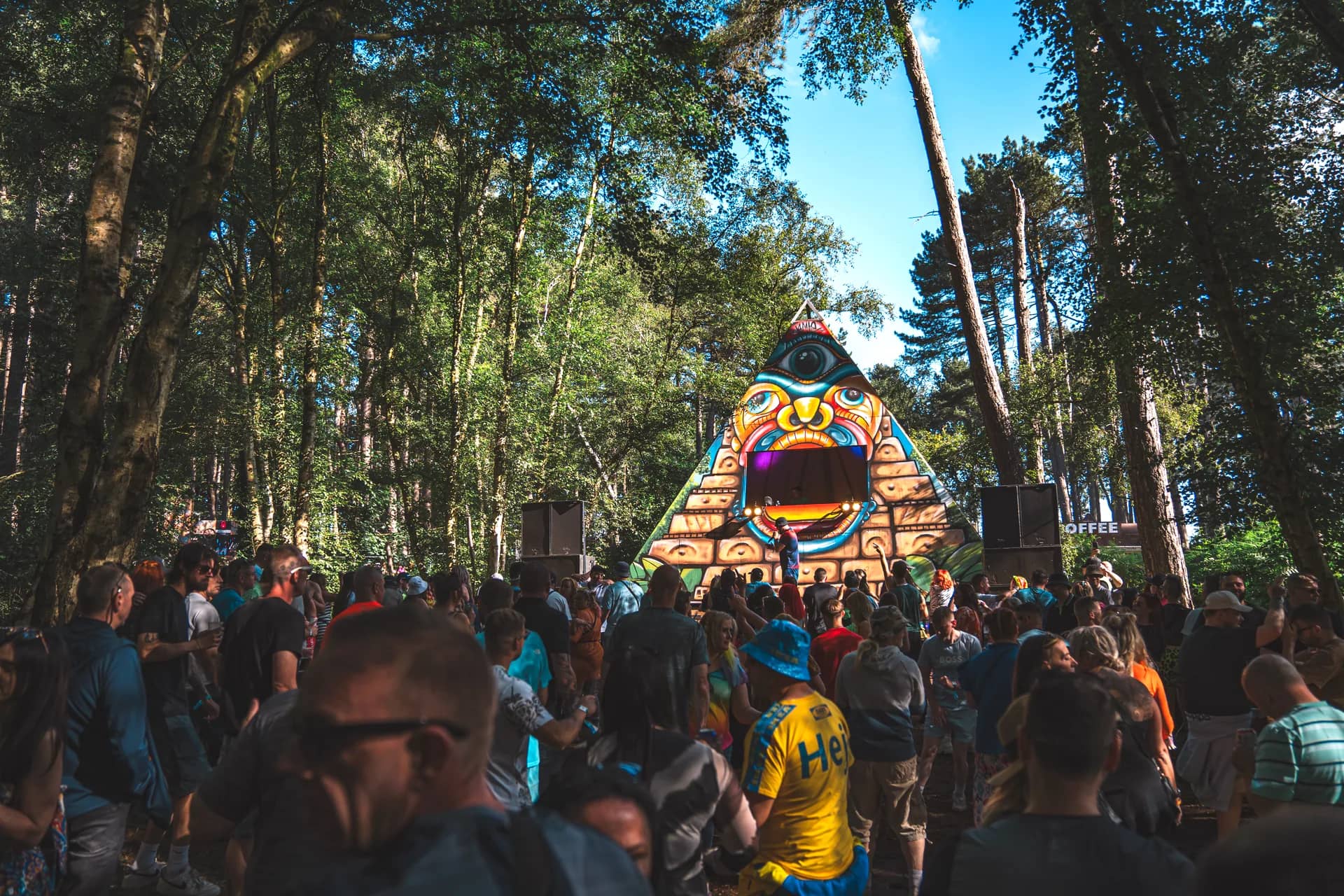 The painted pyramid stage seen through the pines with the crowd gathered in front.