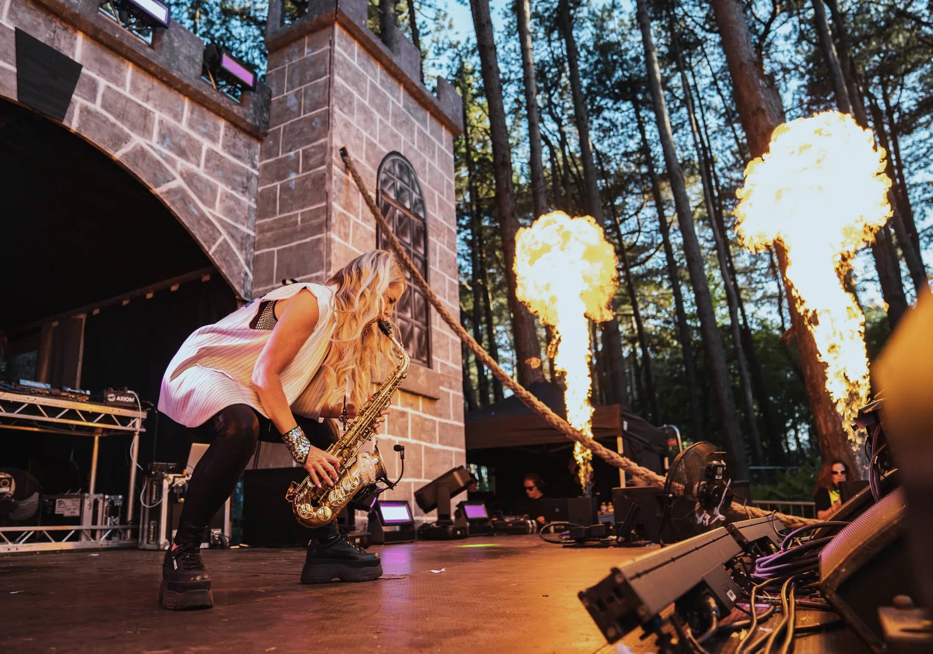 A saxophonist on the castle stage with flame jets firing behind her against the pines.