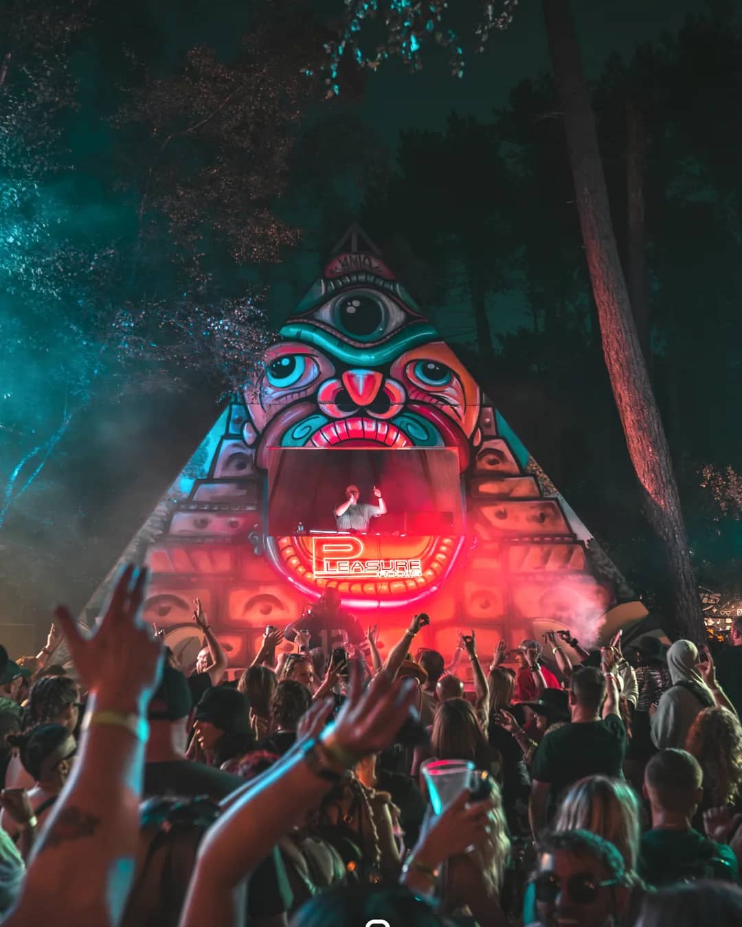The pyramid stage lit teal and pink, a DJ set beneath the eye mural, crowd cheering below.