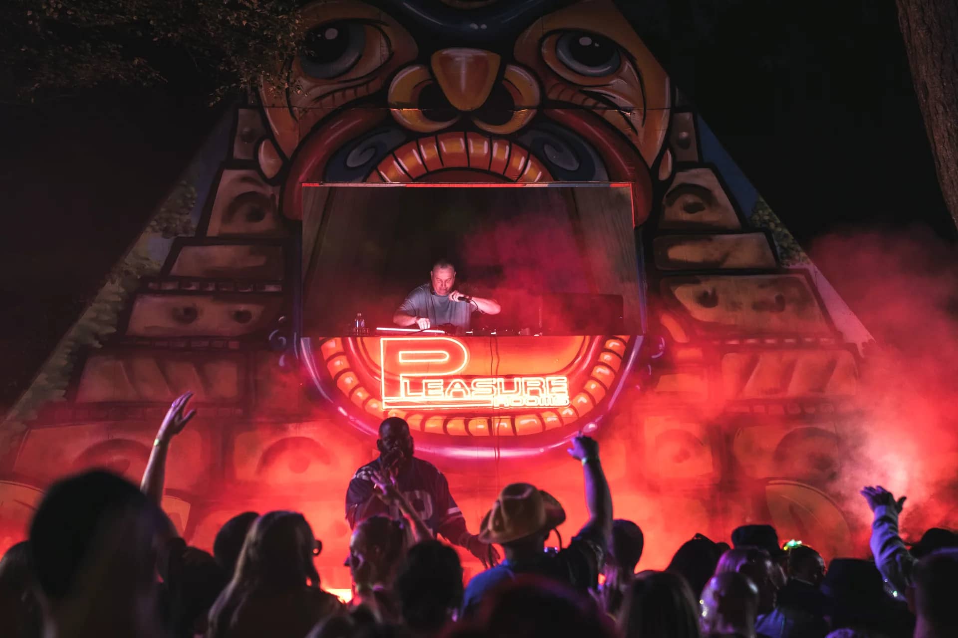 A DJ set inside the pyramid stage's mouth, a red neon Pleasure Rooms sign lighting the crowd below.
