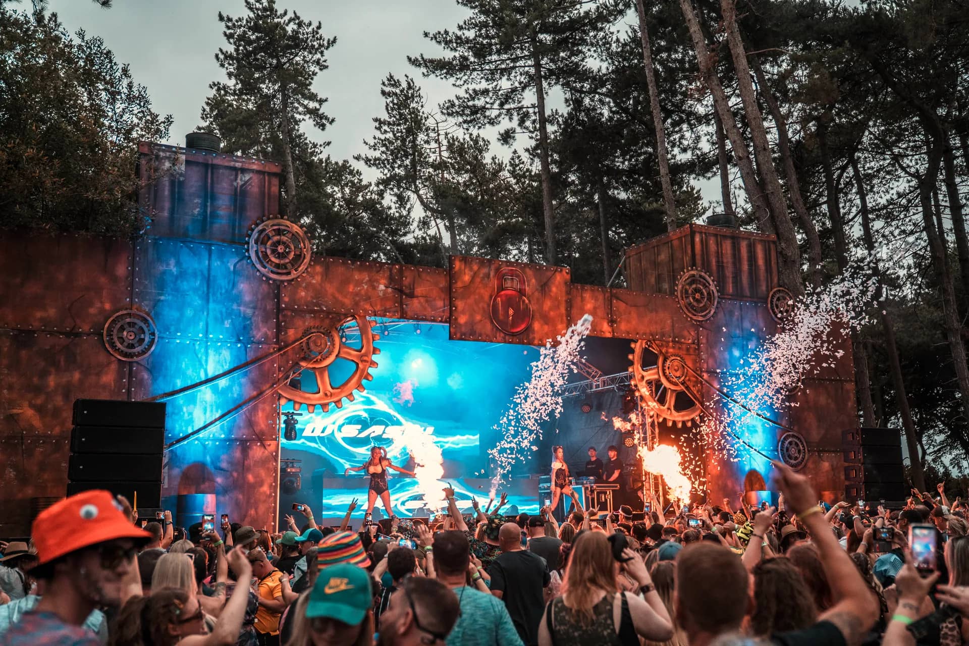 A DJ performing on the steampunk gear stage, fire jets either side and confetti falling over the crowd.