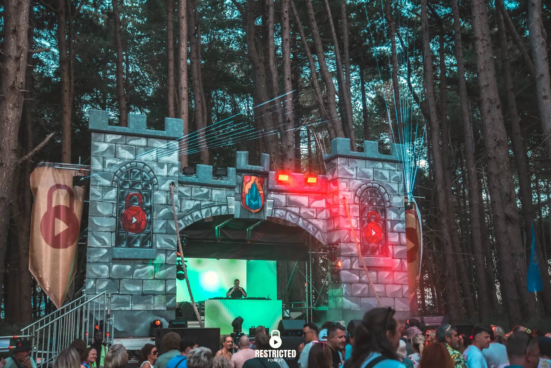 The stone castle gatehouse at dusk: two crenellated towers, the arched windows carrying the padlock crest, banners hanging either side and lasers fanning out over the crowd.