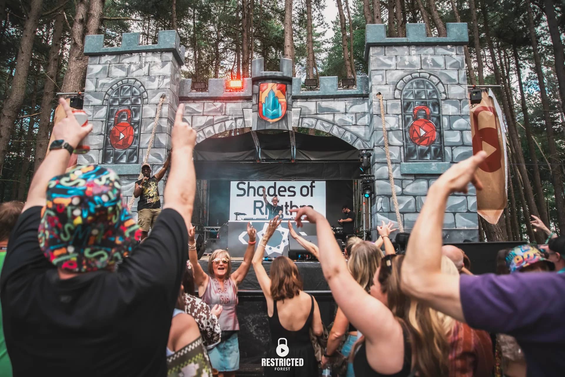 Hands raised in front of the Castle stage in daylight, the DJ's name on the screen inside the arch. Source frame RestrictedForest2026-1329.