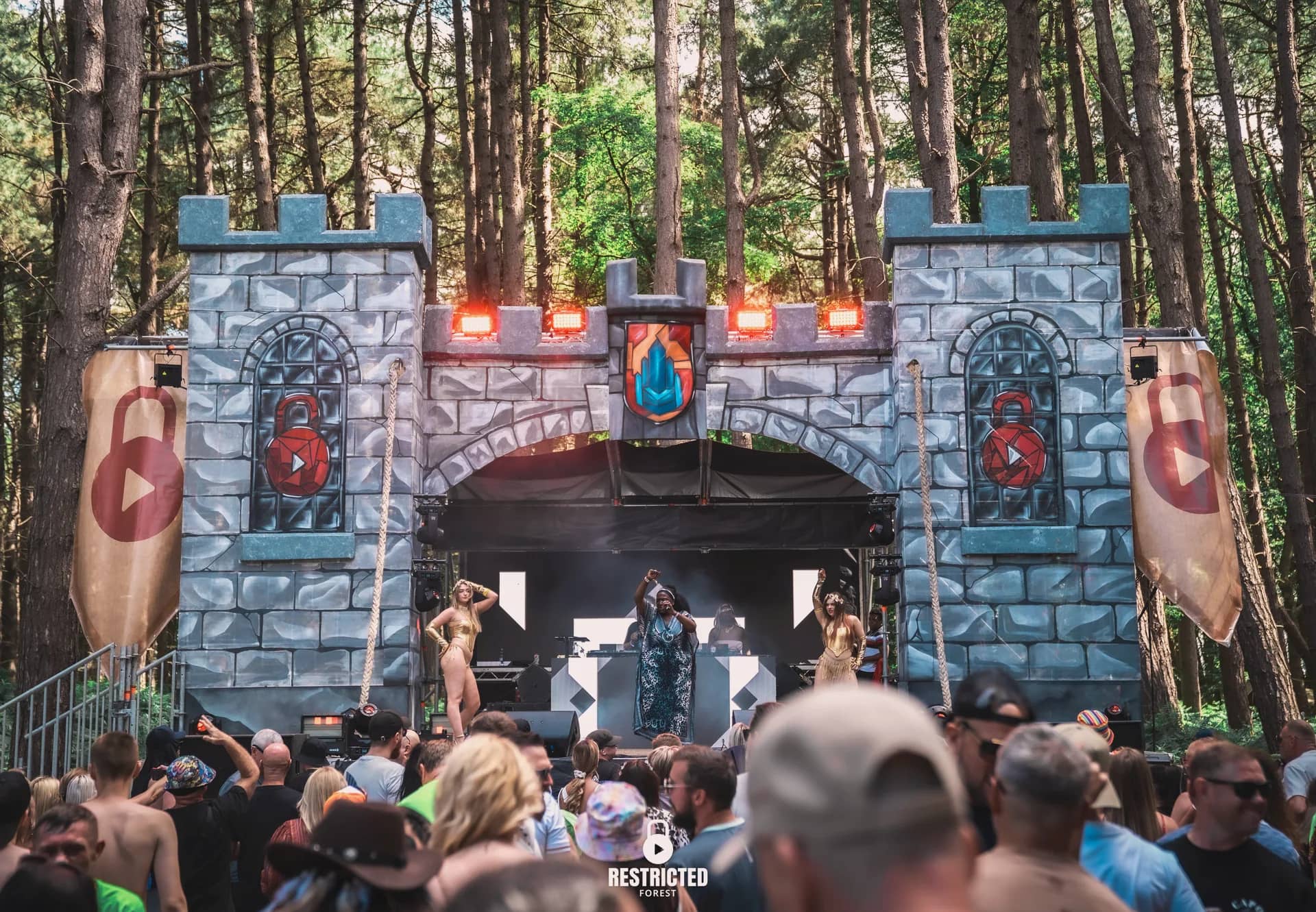 Performers on the Castle stage deck in daylight, the crowd close in front of the arch. Source frame RestrictedForest2026-1140.