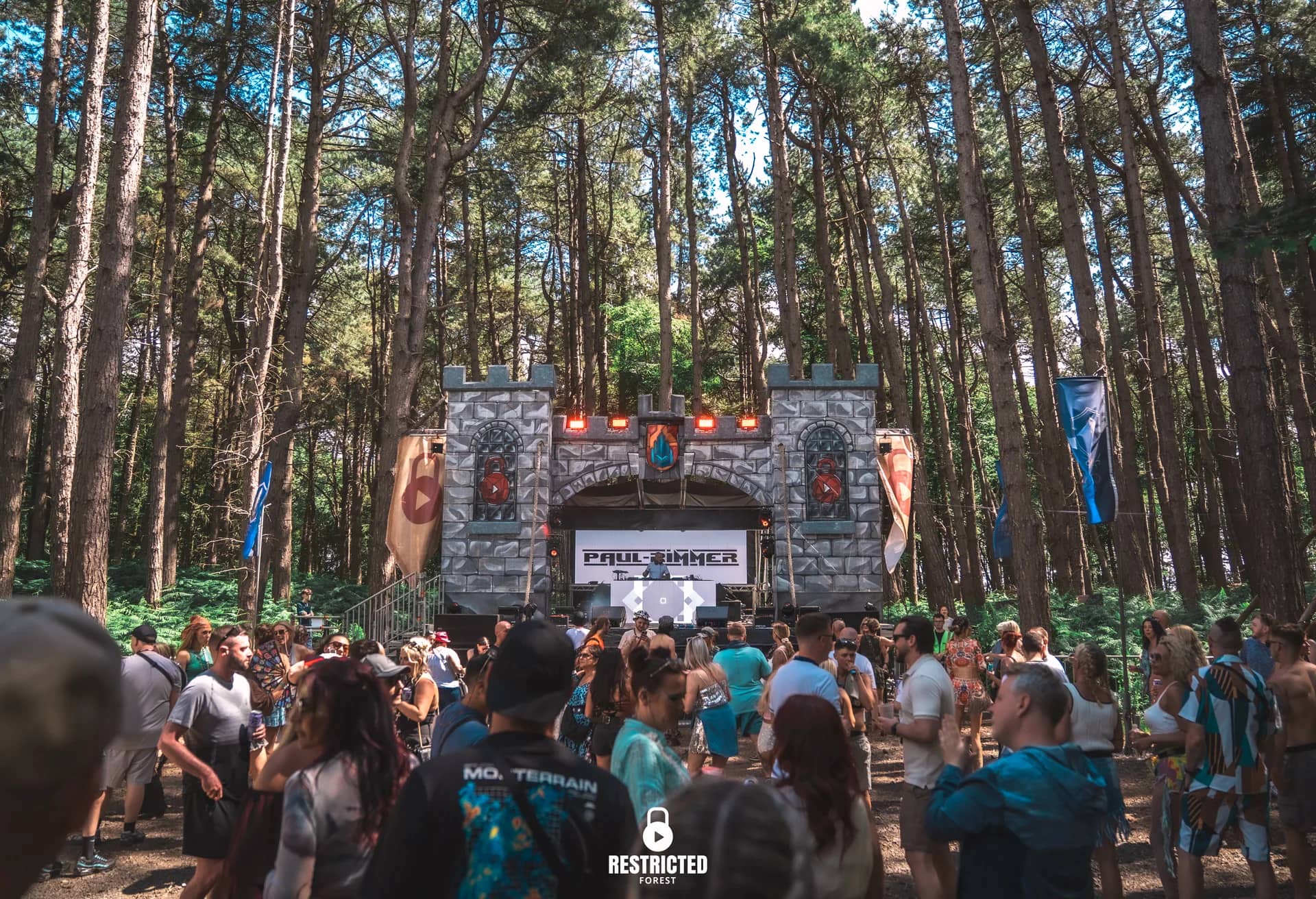 The Castle stage from the back of the clearing in daylight, the grey gatehouse between the pines, banners either side and the crowd spread in front. Source frame RestrictedForest2026-1096.