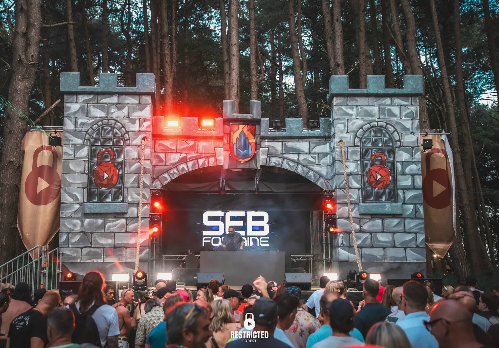 The Castle stage at dusk, red light and smoke inside the arch, the crowd silhouetted in front. Source frame RestrictedForest2026-1701.