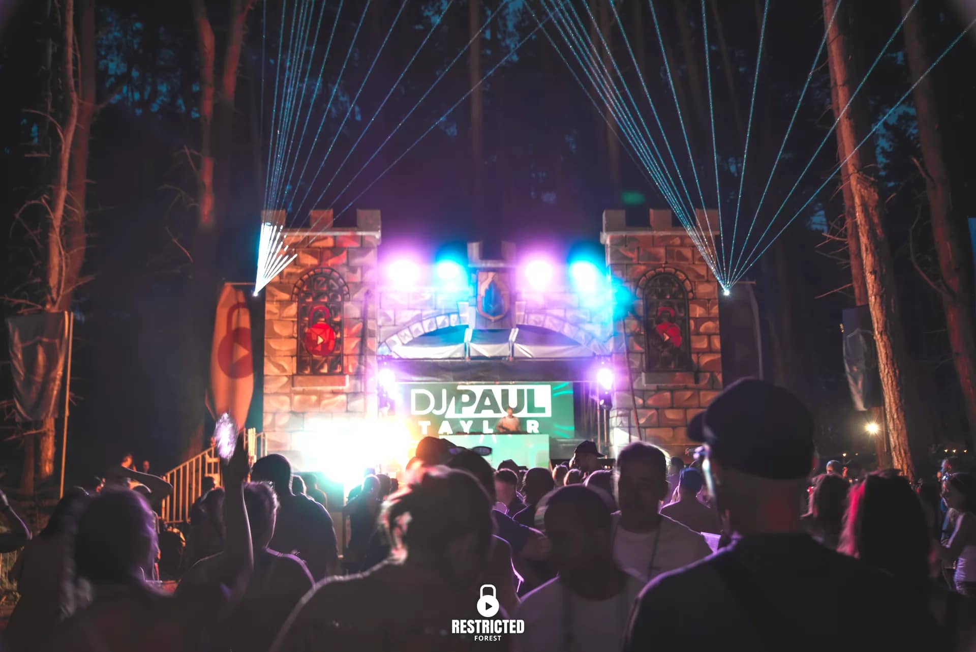 The Castle stage at night, lasers fanning from the towers, the DJ's name lit on the screen. Source frame RestrictedForest2026-1950.