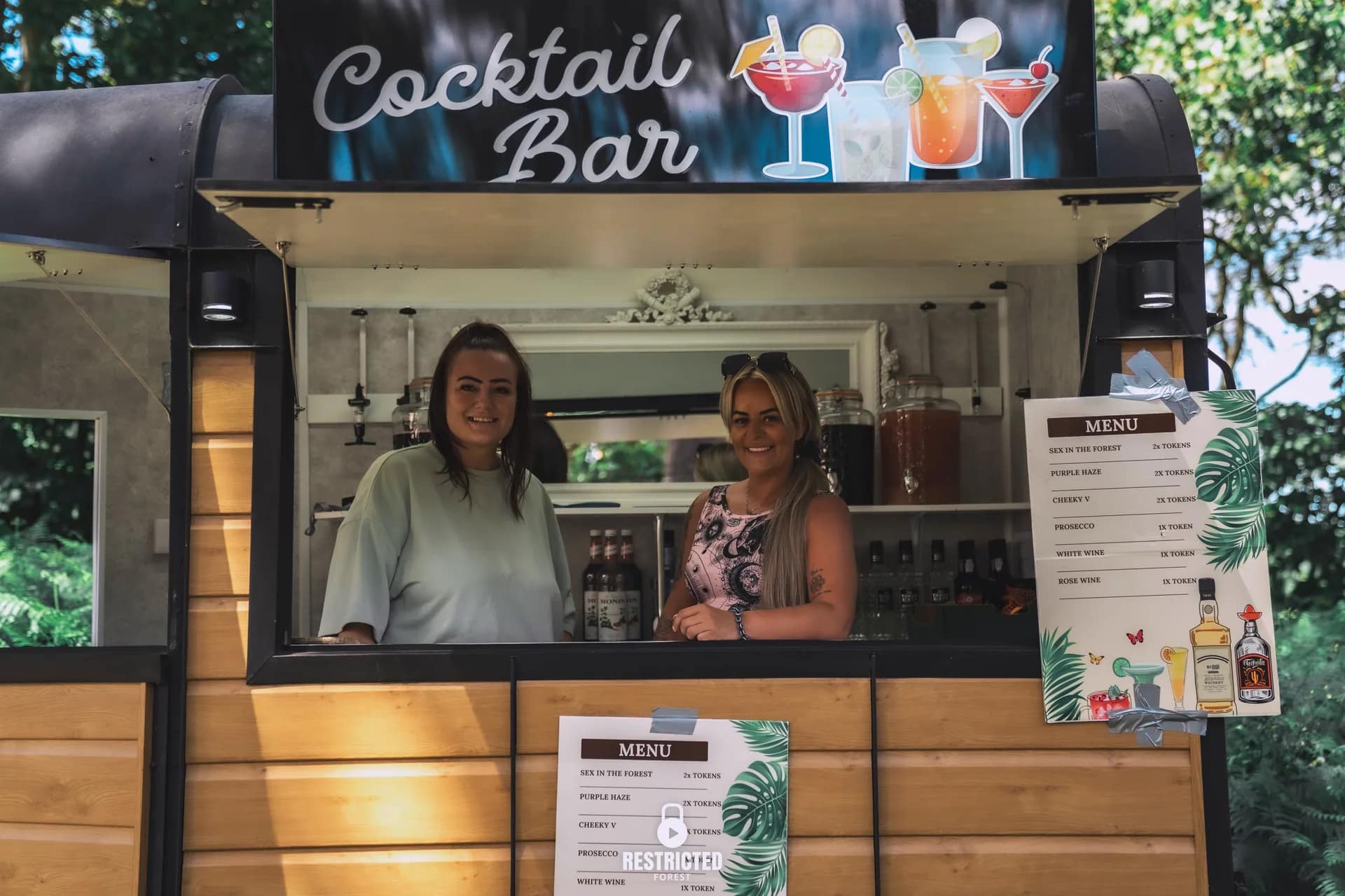 Two bar staff behind the counter of the Cocktail Bar kiosk in the trees, menu boards either side of them. Source frame RestrictedForest2026-1010.