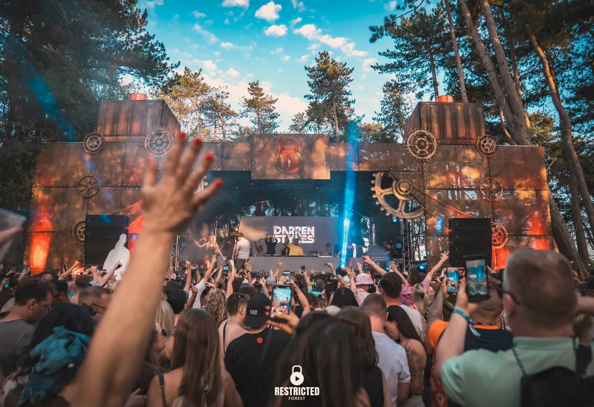 Darren Styles on the factory gear stage in daylight, crowd with hands raised and phones out, a laser beam cutting through the smoke.