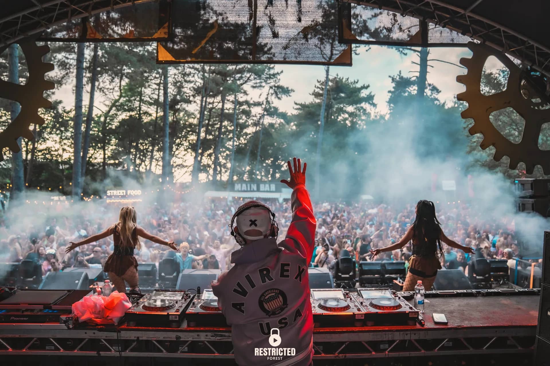 Two dancers with arms outstretched either side of the DJ on the factory stage, confetti falling over the crowd.