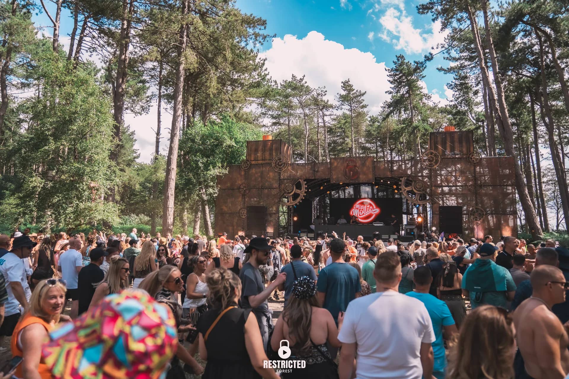 The Factory stage from the back of the clearing in daylight, the whole cog-and-girder build under the pines, crowd stretching forward. Source frame RestrictedForest2026-1112.