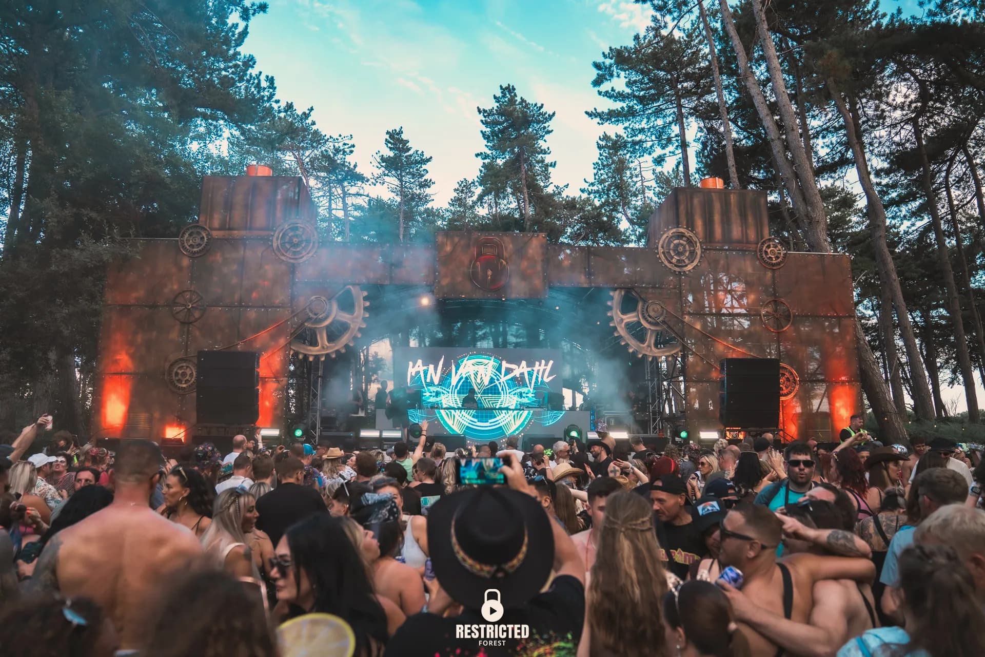 The cog and girder stage lit through heavy haze, crowd stretching back into the woods.