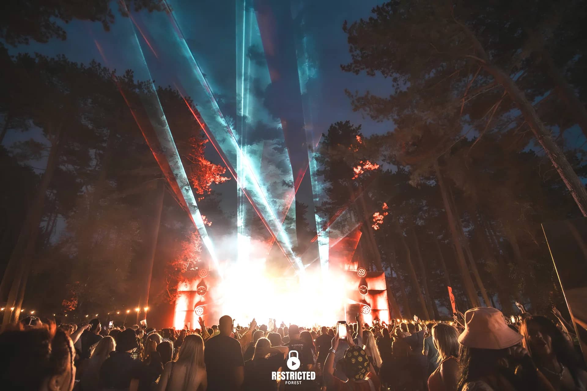 Lasers fanning up through the pines from the Factory stage at night, the crowd lit red and blue below. Source frame RestrictedForest2026-1947.