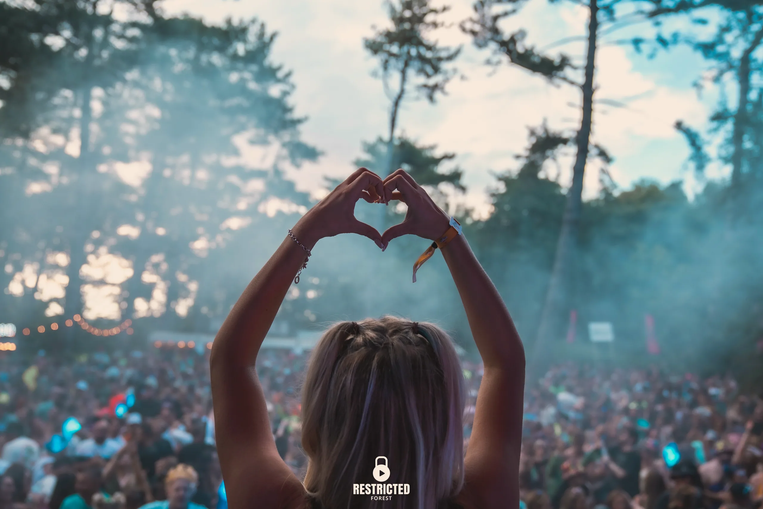 Someone at the front of the crowd making a heart with both hands, the field and the treeline in haze beyond. Source frame RestrictedForest2026-1603.
