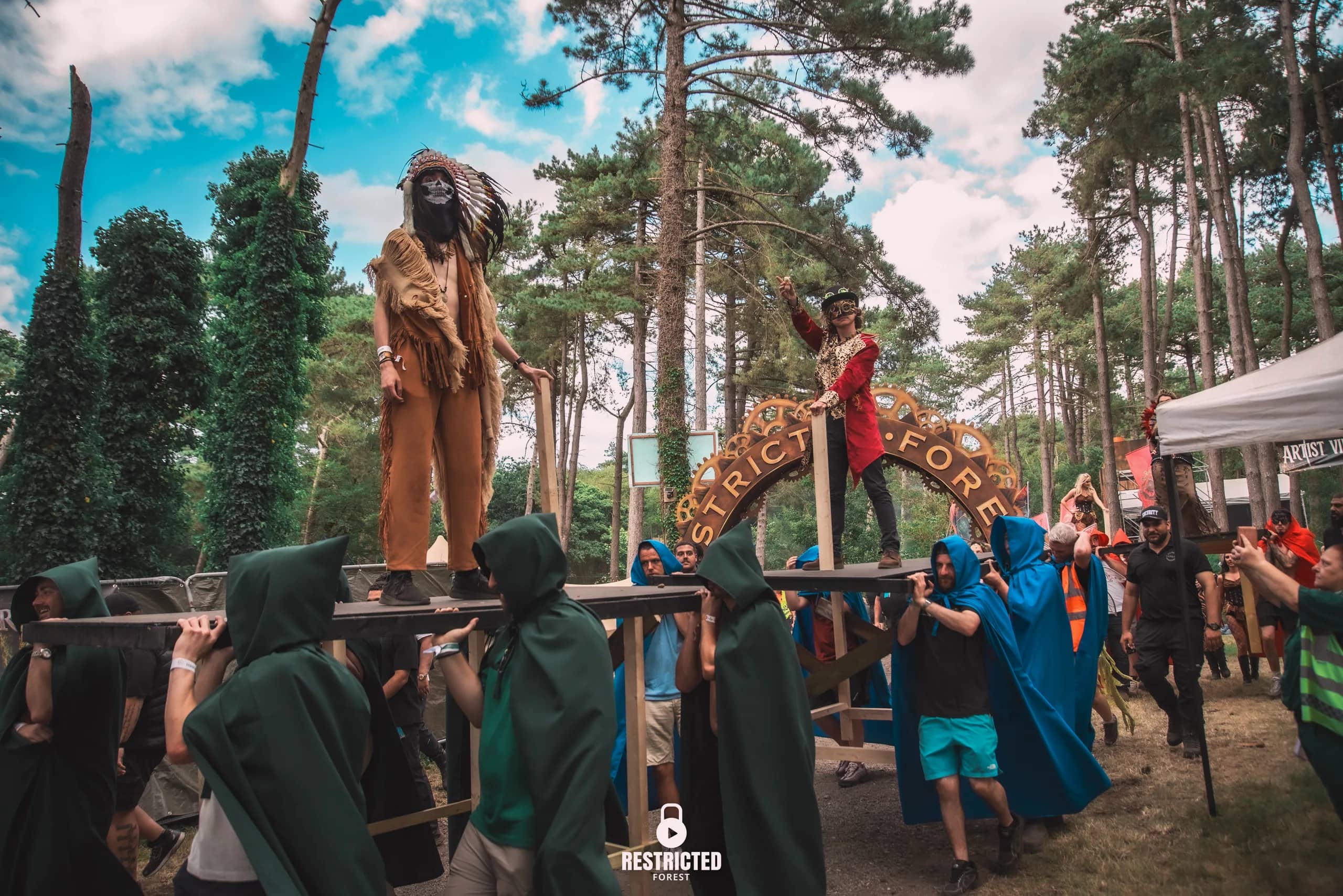 The procession: two figures carried above the crowd on platforms by green and blue cloaked bearers, the cog arch behind. Source frame RestrictedForest2026-1494.