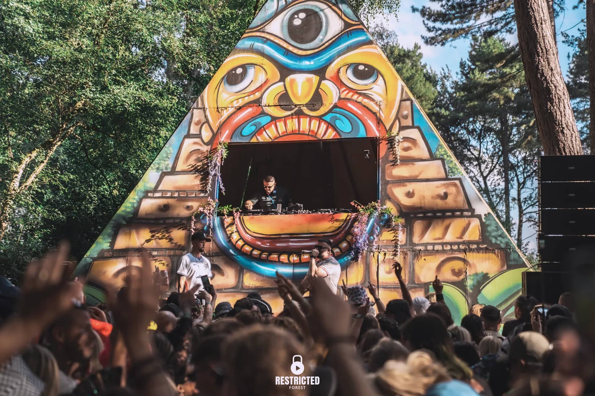 The painted pyramid stage in daylight, hands raised across the crowd below its mouth. Source frame RestrictedForest2026-1500.