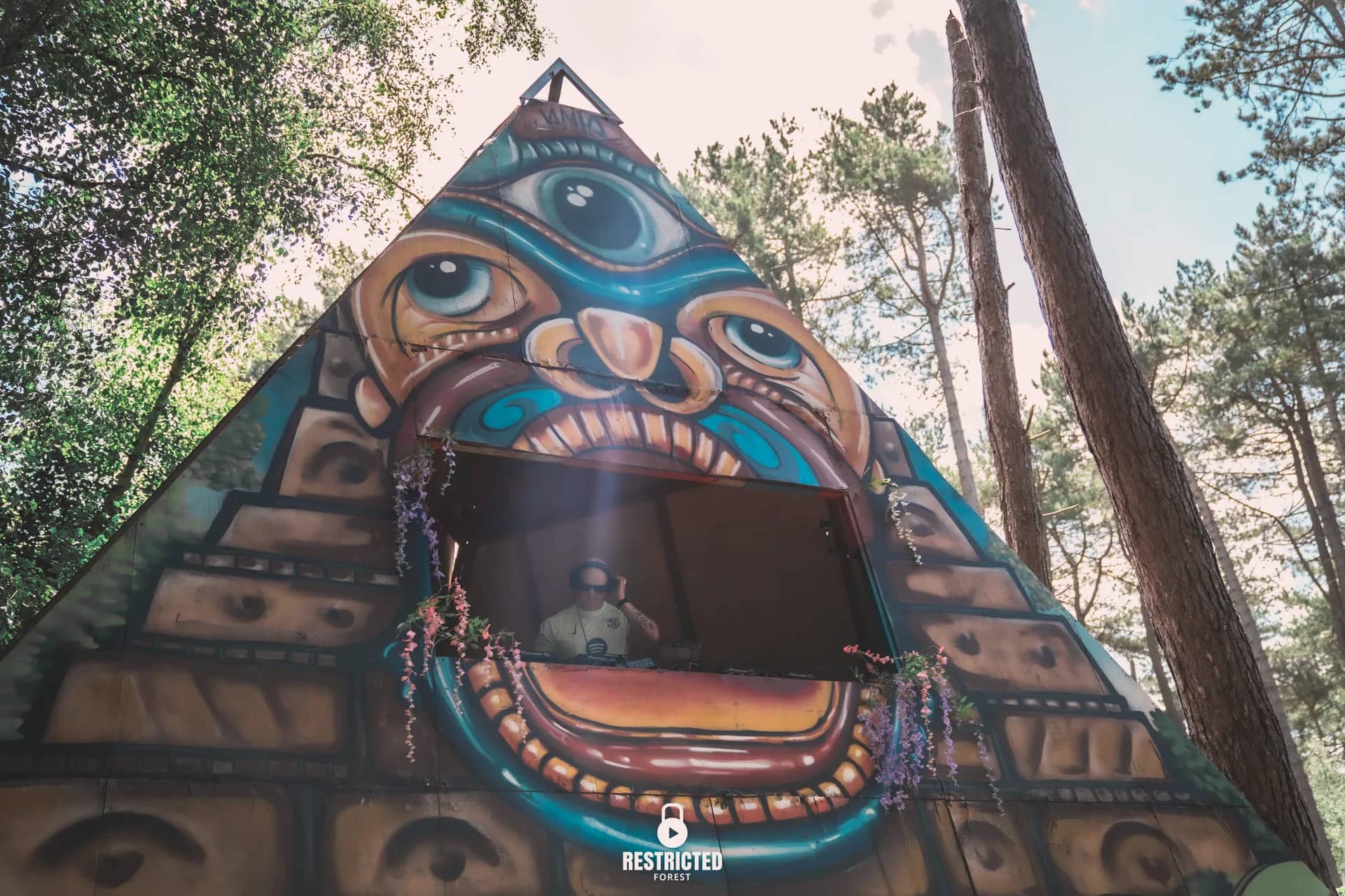 The painted pyramid stage from below in daylight, the DJ in its mouth under the eye. Source frame RestrictedForest2026-1042.