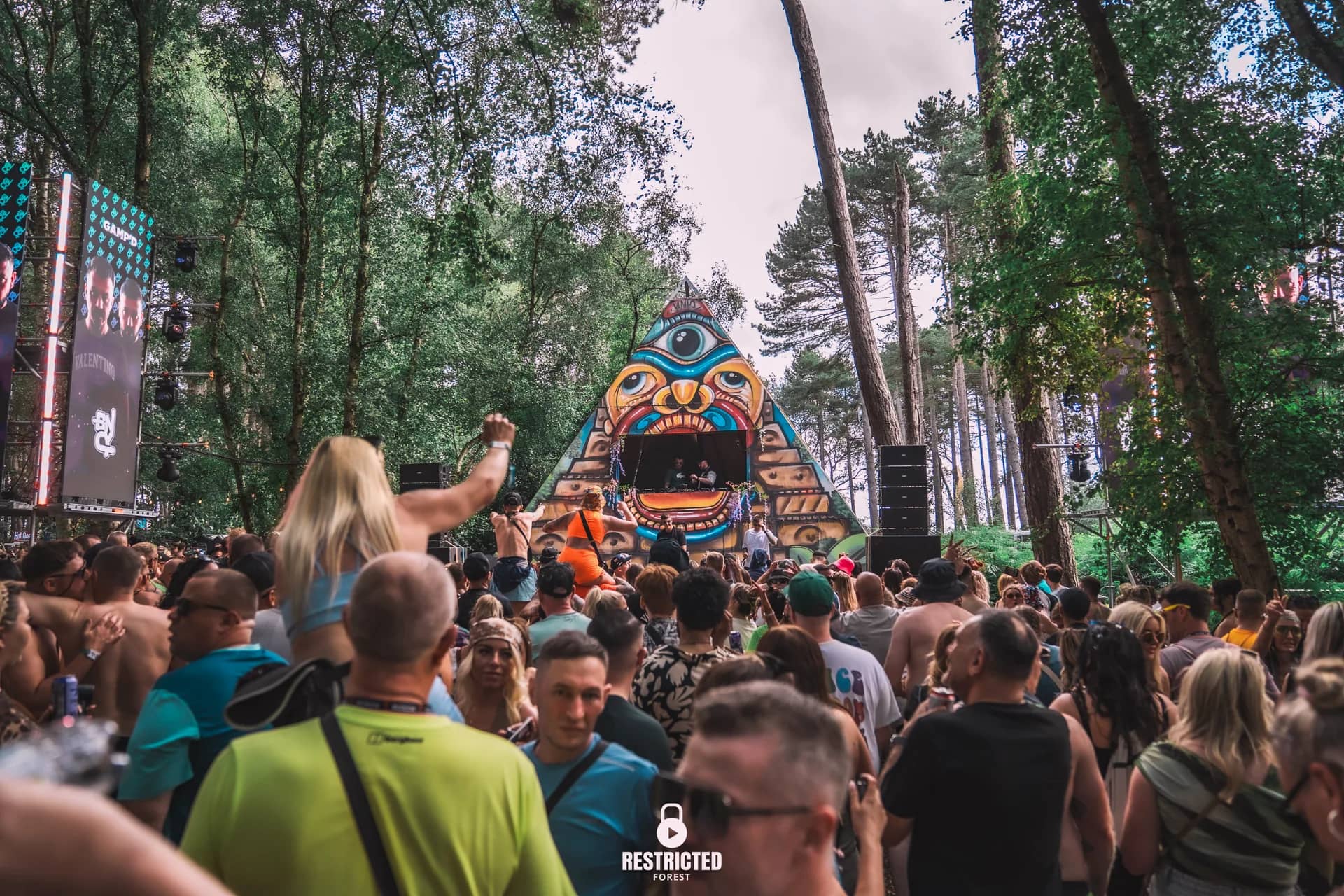 The painted pyramid stage at the end of the clearing in daylight, the crowd filling the path to it. Source frame RestrictedForest2026-1236.