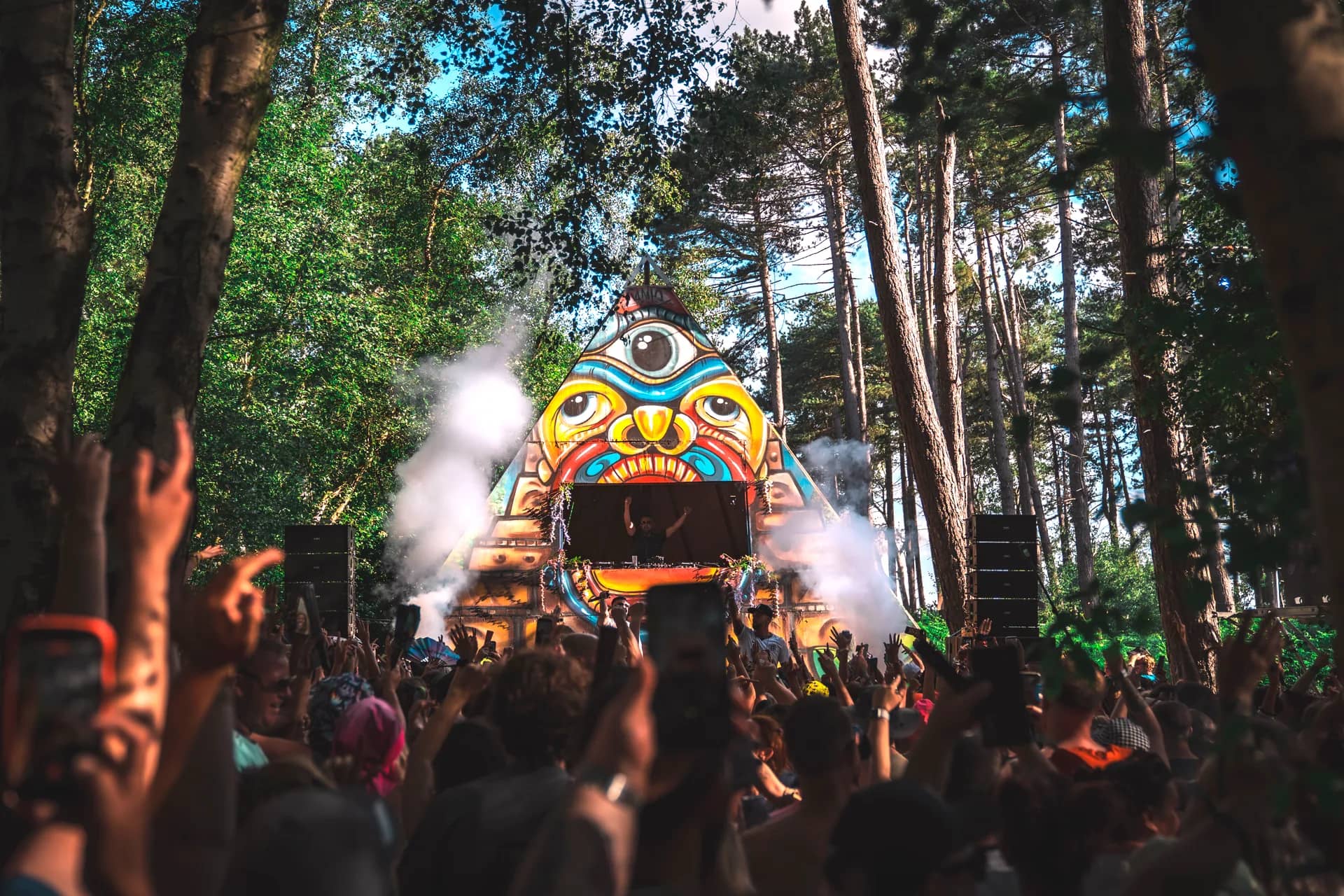 The painted pyramid stage under blue sky, a DJ performing inside its mouth, crowd with arms raised in daylight.