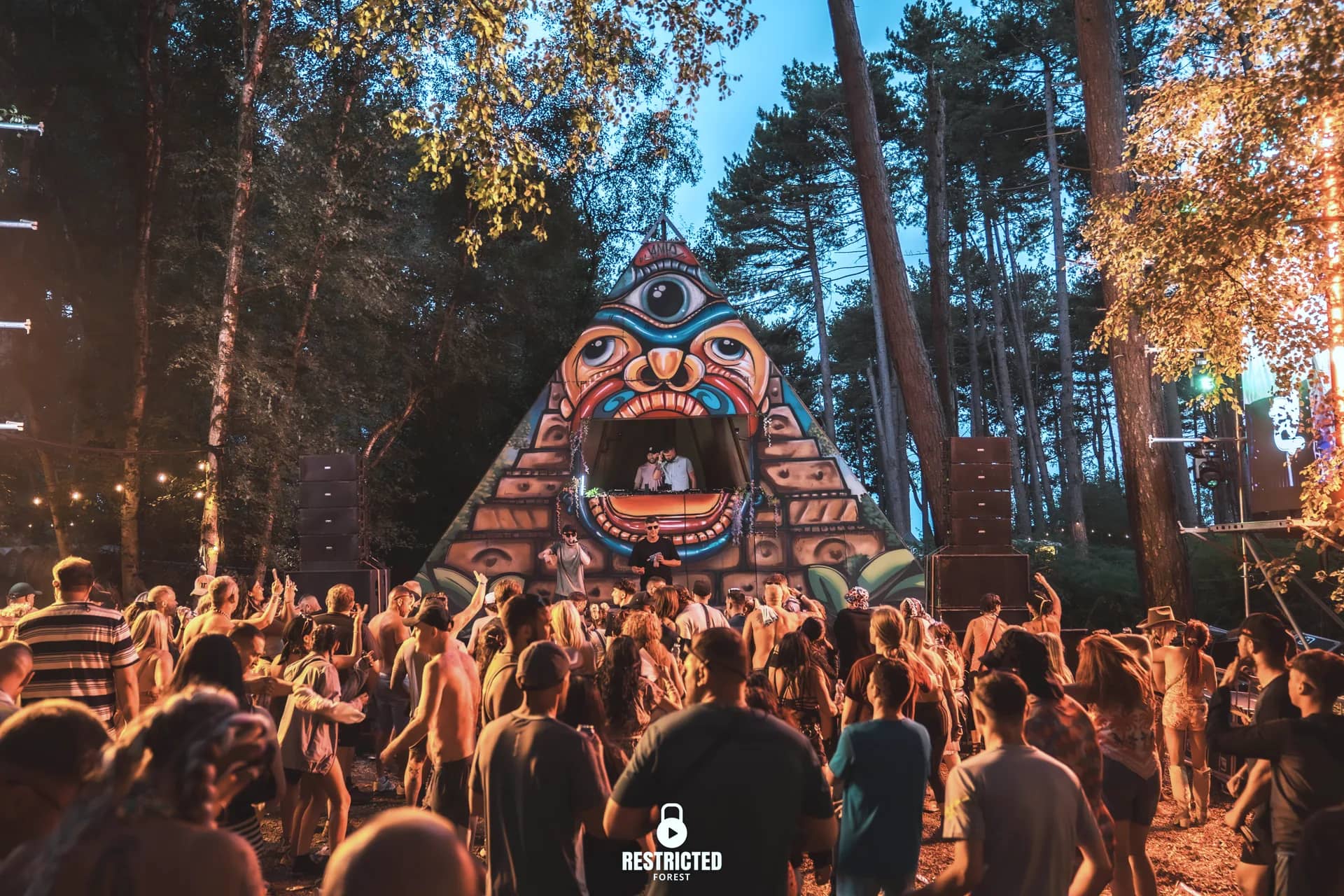 The painted pyramid stage at dusk, the clearing lit amber and the crowd facing it. Source frame RestrictedForest2026-1904.