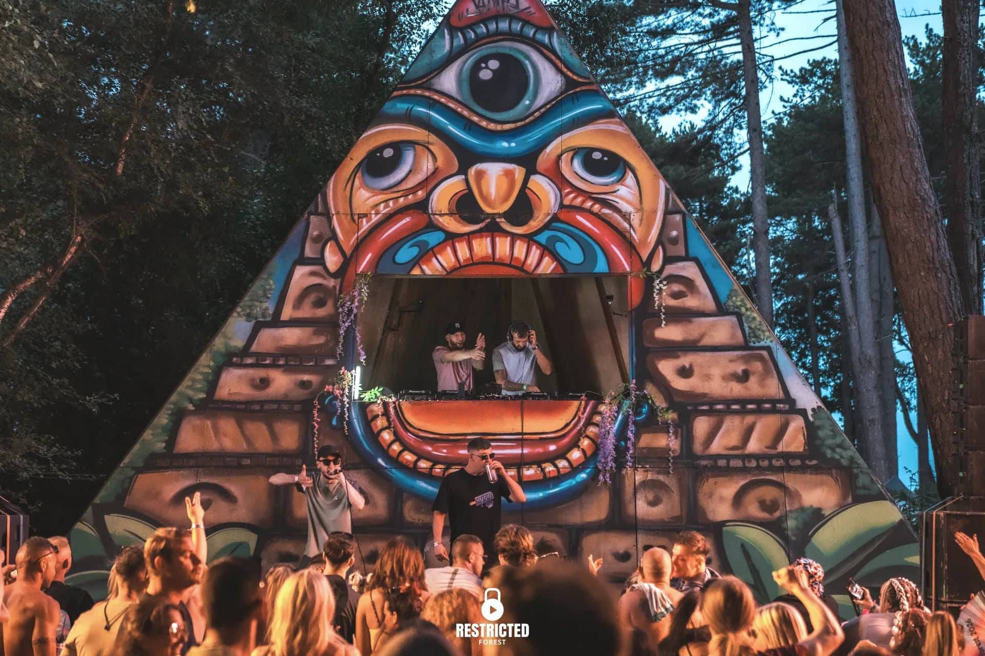 Performers in front of the painted pyramid stage at dusk, the crowd lit warm below. Source frame RestrictedForest2026-1902.