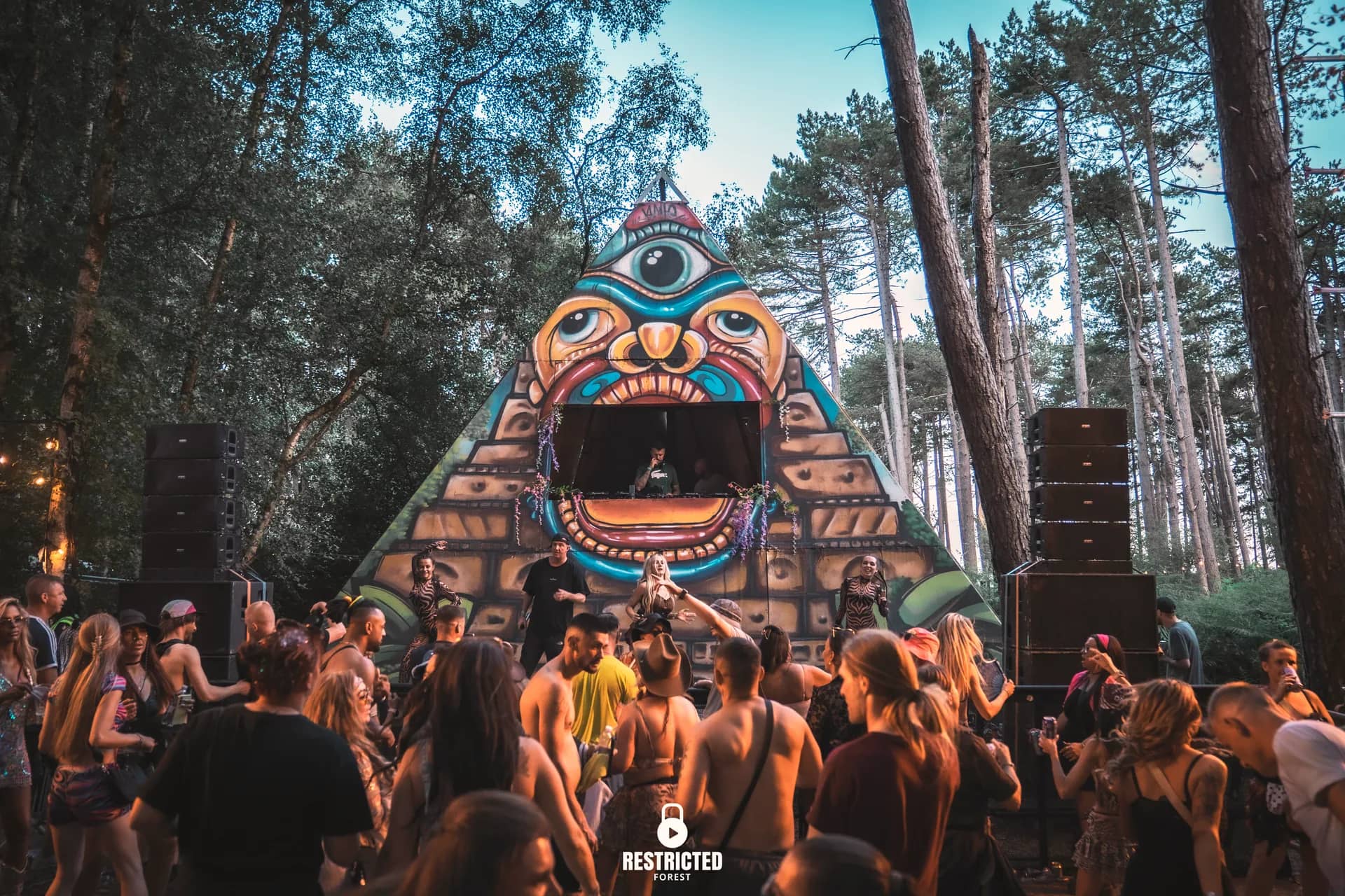The painted pyramid stage in the evening light, the crowd dancing in front and the pines catching the last sun. Source frame RestrictedForest2026-1841.