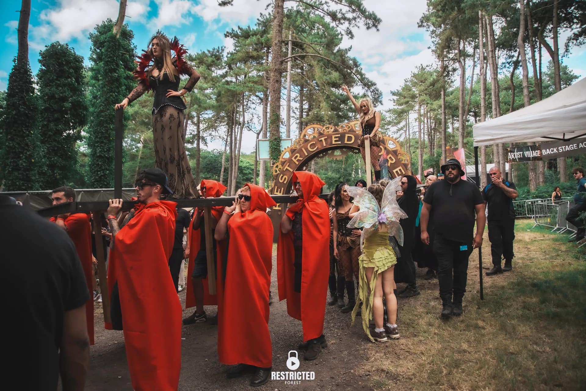 Performers in red hooded cloaks carrying a raised platform through the woods, two costumed performers standing on it, passing under the Restricted Forest arch