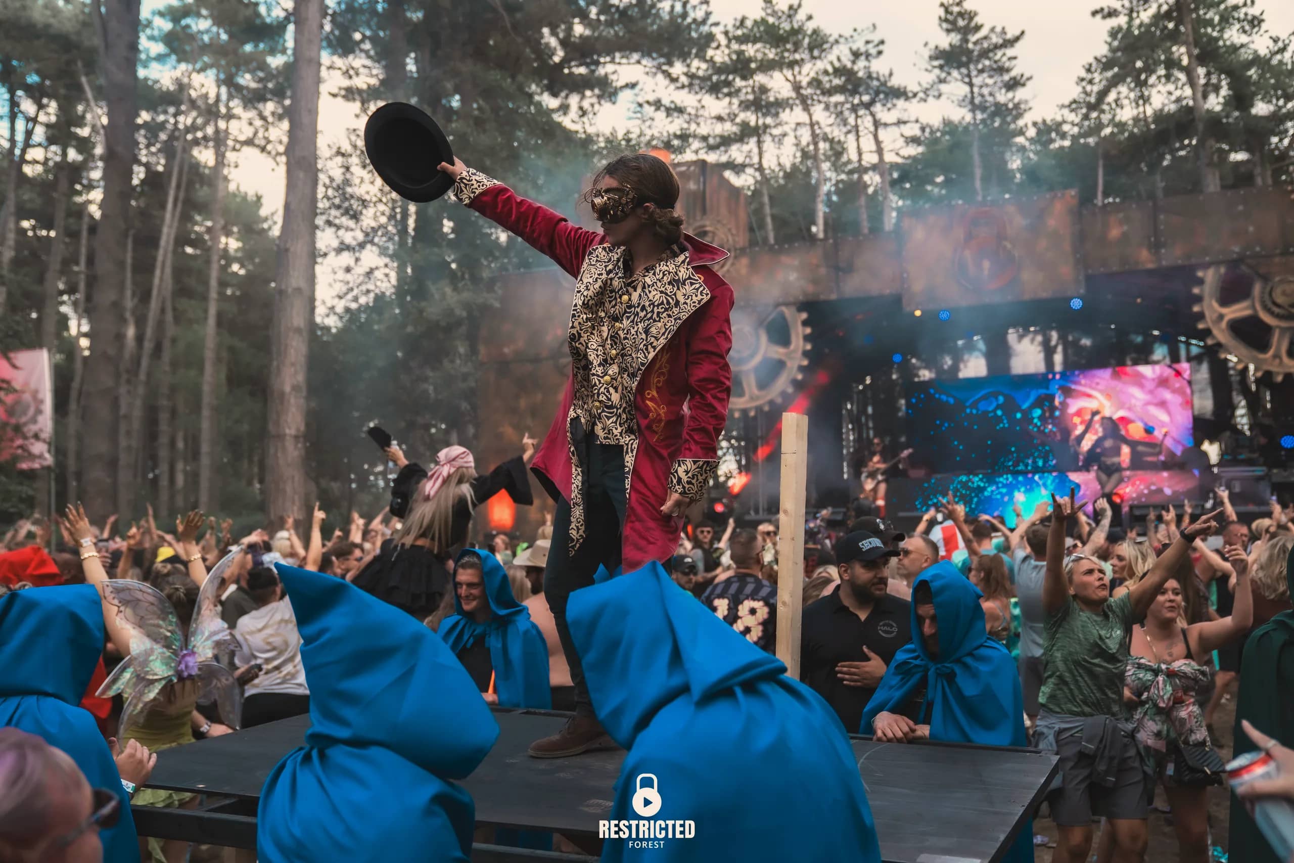 The performer in the red tailcoat raised above the crowd with a top hat held out, the Factory stage and its pyro behind. Source frame RestrictedForest2026-1437.