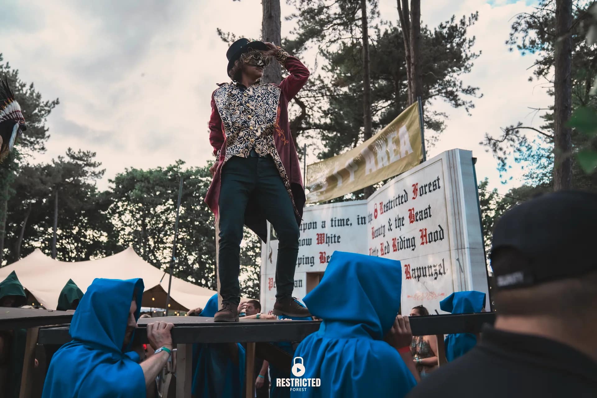 The performer in the red tailcoat standing on a platform by the fairytale gate, blue-cloaked bearers below. Source frame RestrictedForest2026-1423.