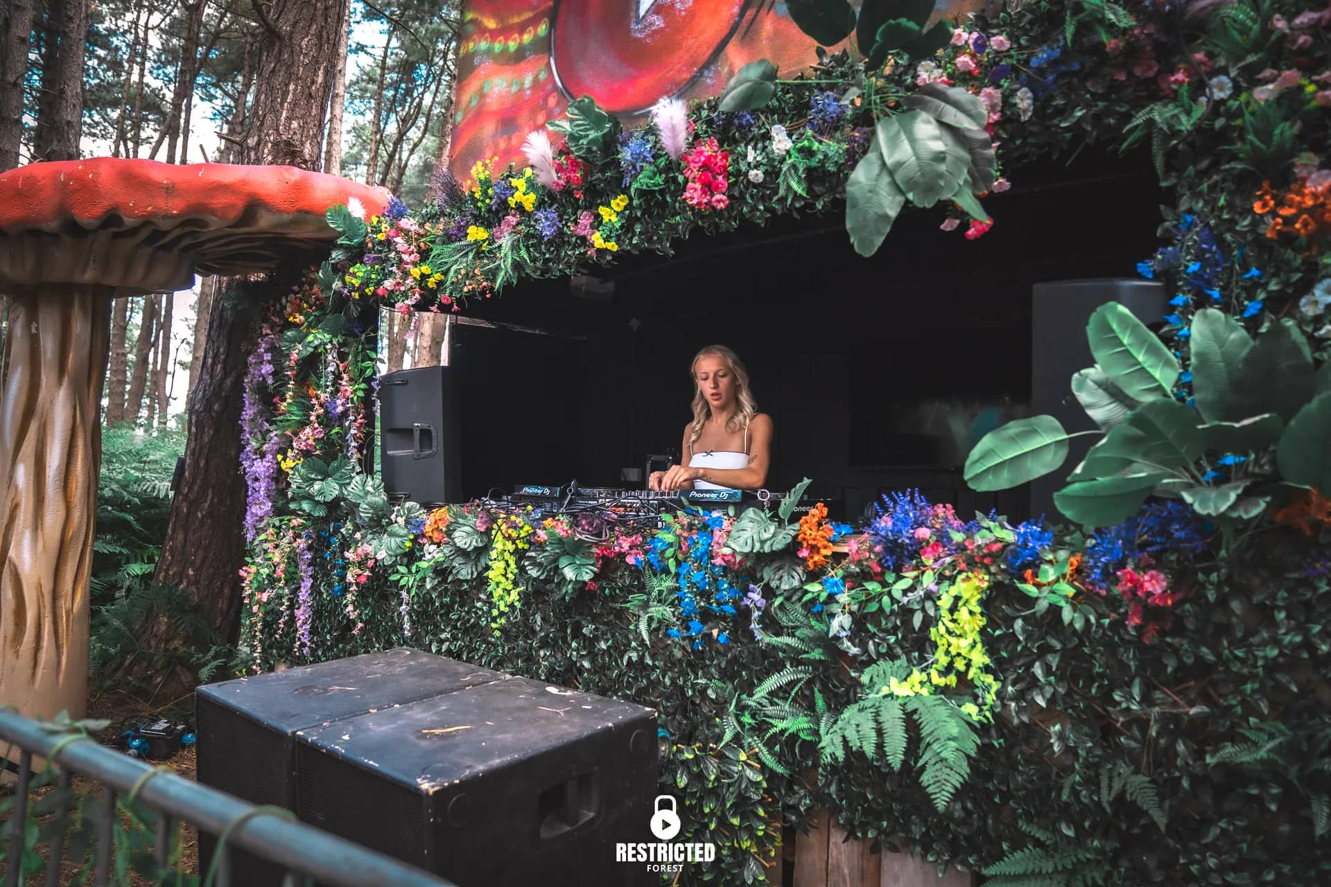 The Secret Garden's flower-draped booth beside a giant red toadstool, the DJ small inside it. Source frame RestrictedForest2026-1036.