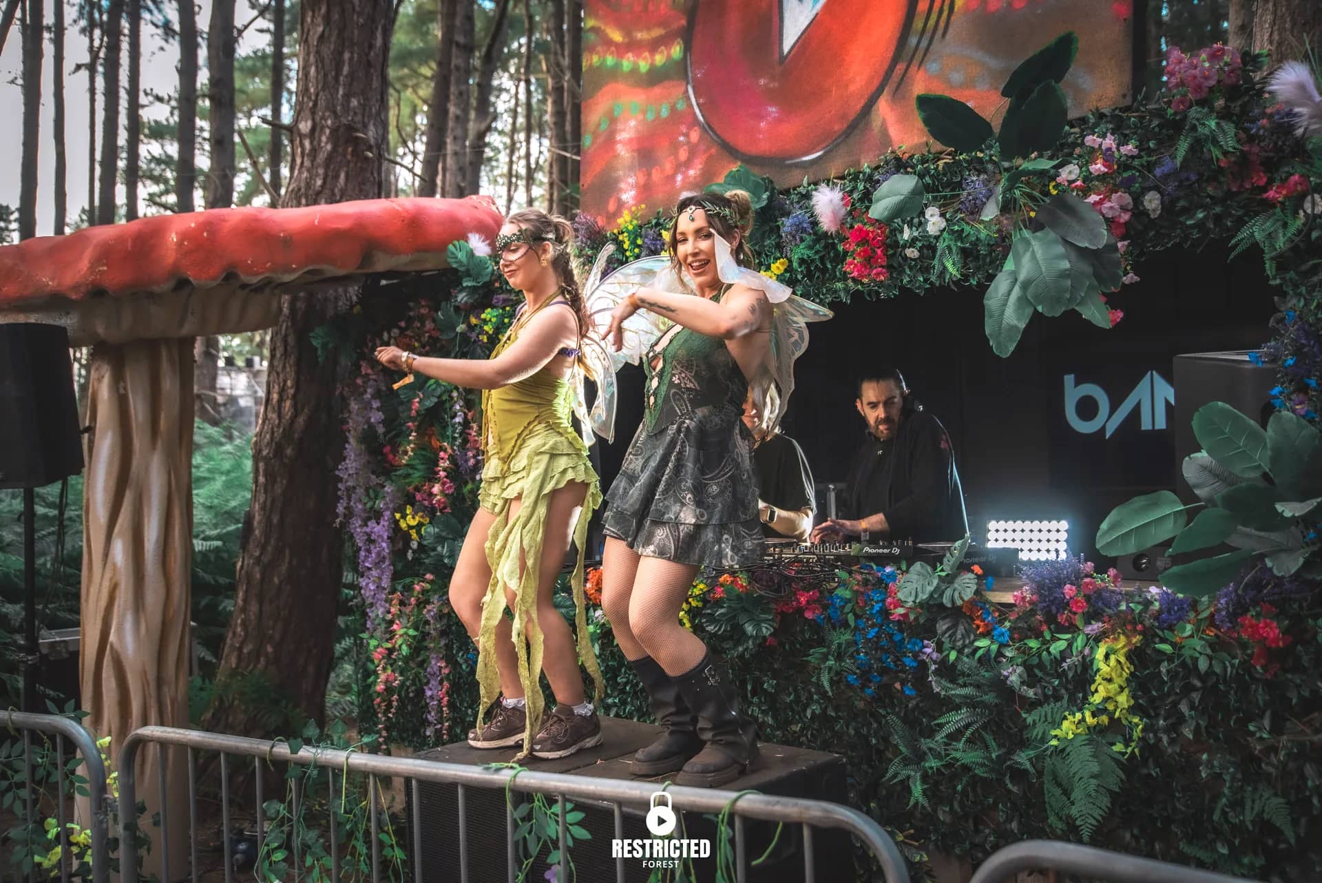 Two fairy performers on the Secret Garden booth's rail beside a giant toadstool, the DJ behind them. Source frame RestrictedForest2026-1739.