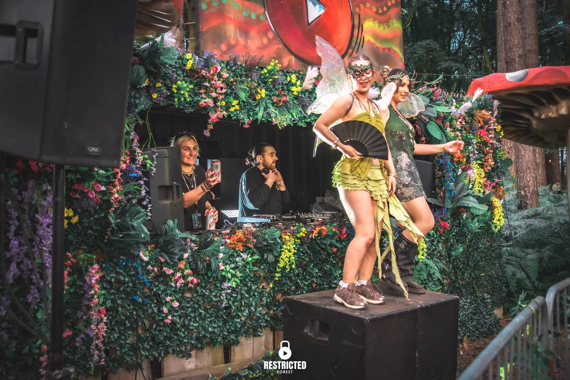A fairy performer with a fan dancing on the Secret Garden booth, flowers and leaves around her. Source frame RestrictedForest2026-1746.