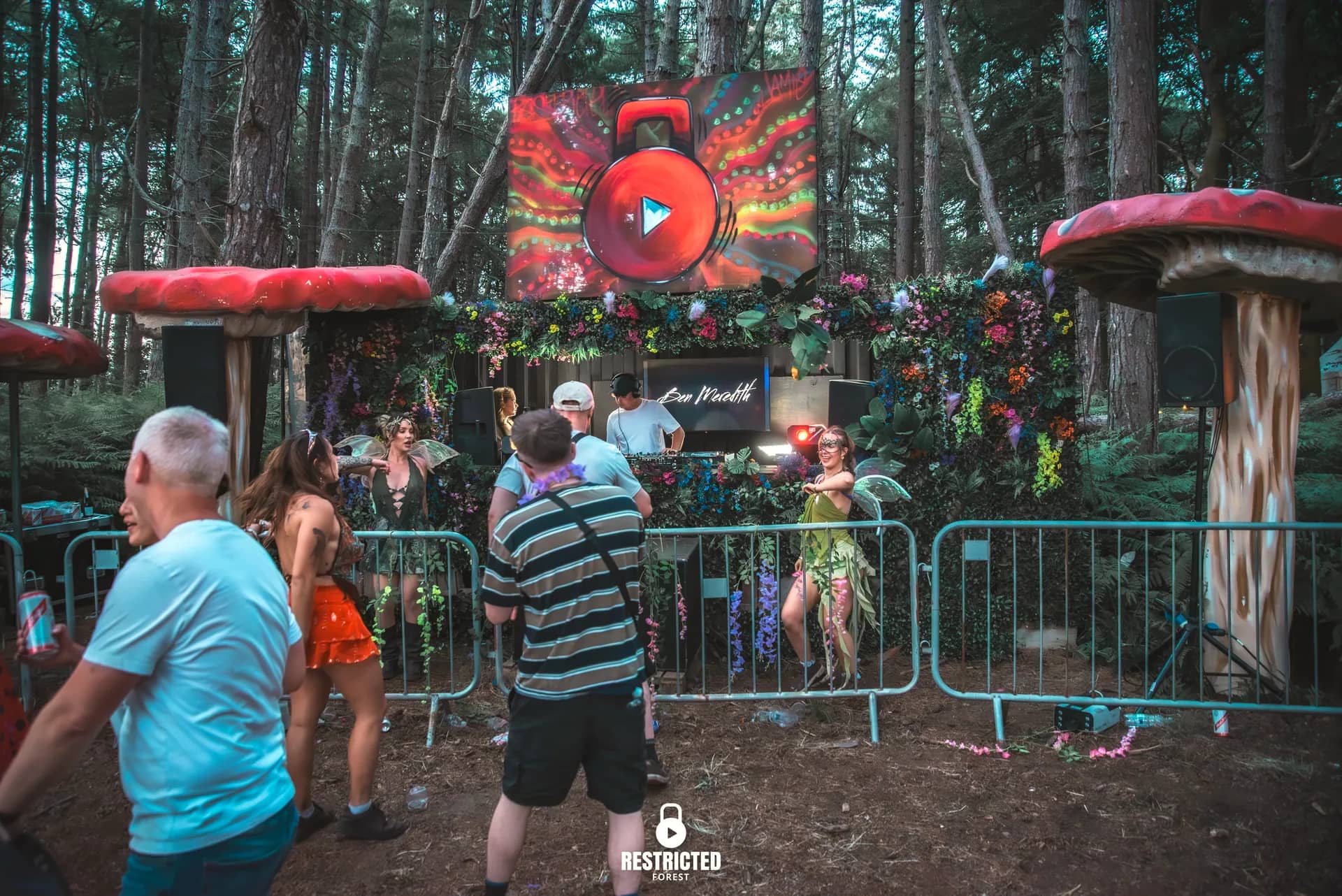 The Secret Garden stage in daylight: the flower-draped booth between two giant toadstools, a fairy performer on the rail and the crowd gathering under the pines. Source frame RestrictedForest2026-1885.