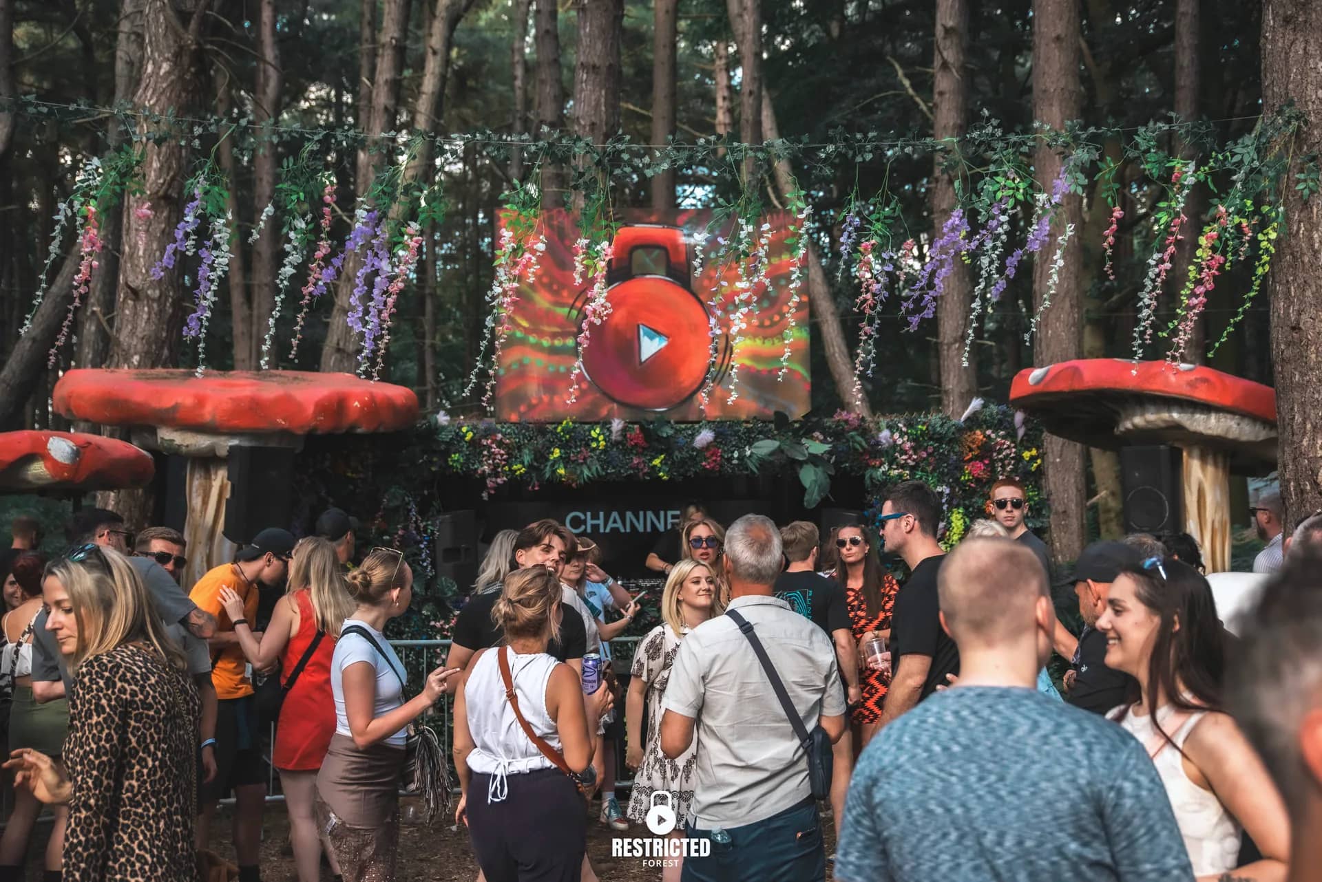 The Secret Garden in daylight, wisteria hung from the trees over the booth and a crowd gathered in front. Source frame RestrictedForest2026-1524.