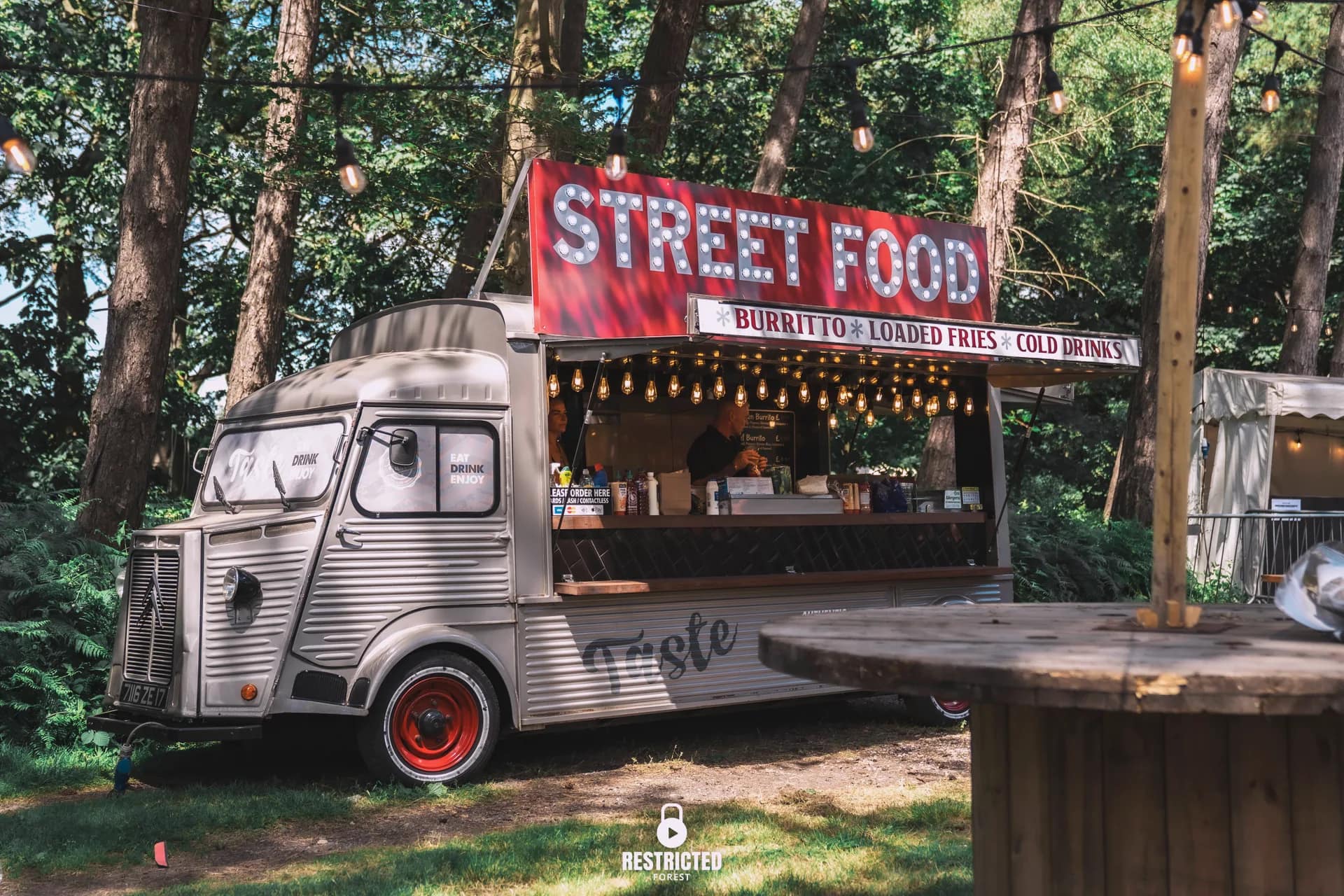 A vintage Citroen H van serving street food under the pines, strung with lights and signed for burritos and loaded fries. Source frame RestrictedForest2026-1009.