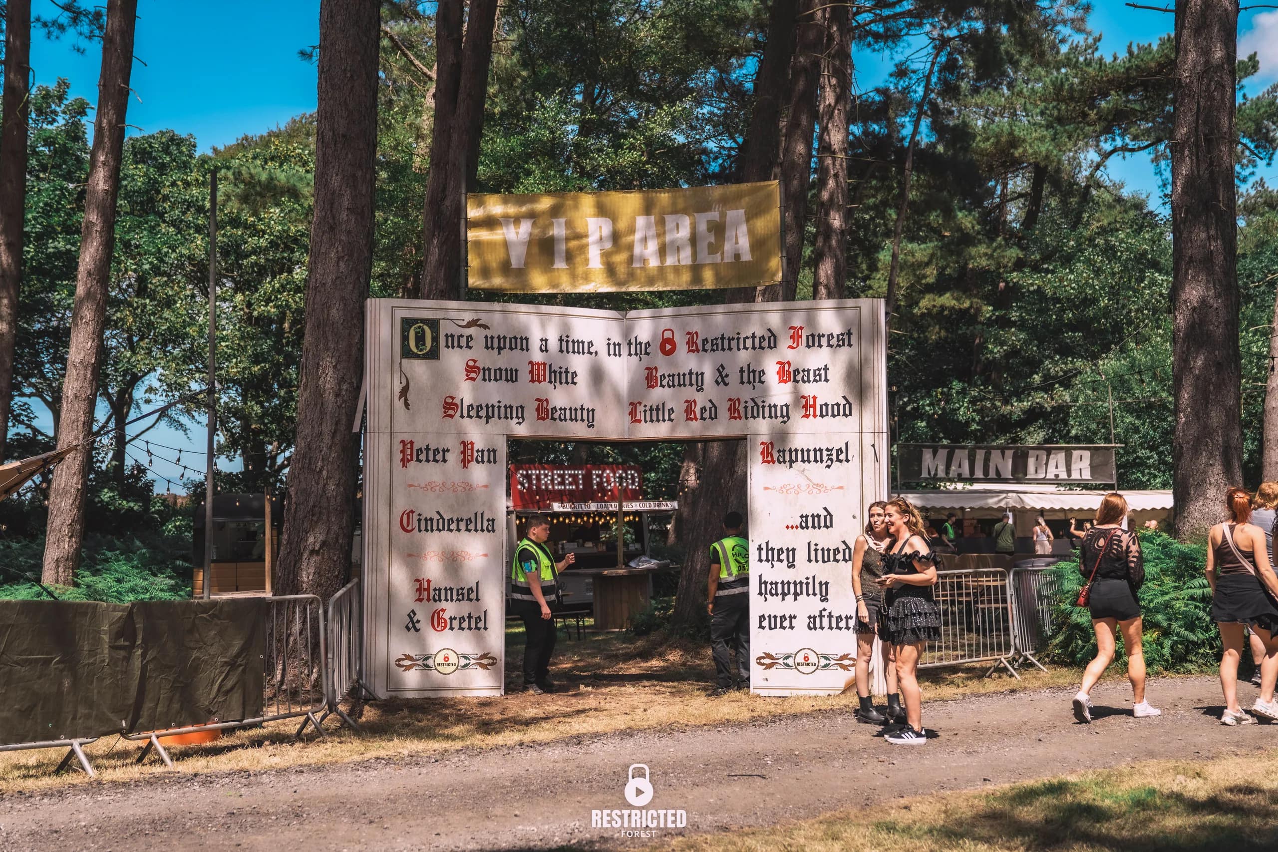 The VIP area gate, its board lettered with fairytale names and ending on happily ever after, the main bar behind. Source frame RestrictedForest2026-1018.