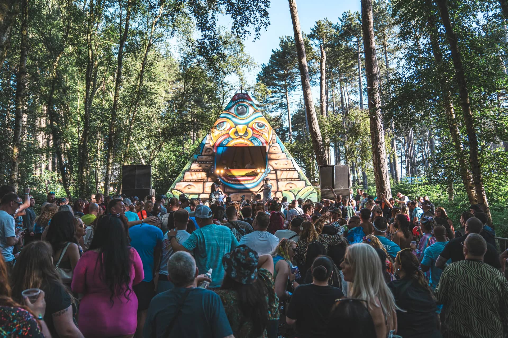 Painted totem mask stage seen over the crowd, tall pines framing it