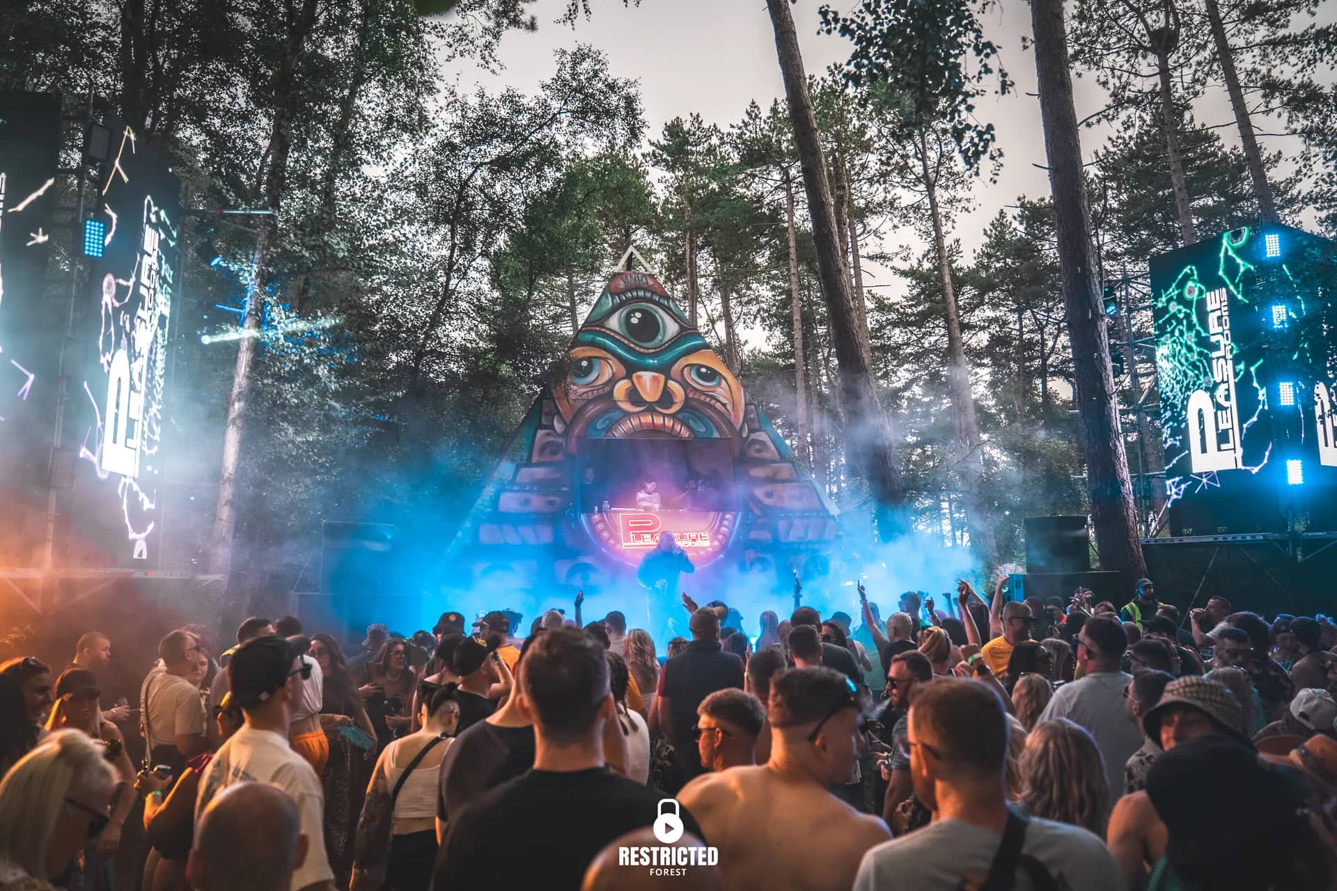 Carved totem stage lit blue at dusk, haze over the crowd, pines behind.